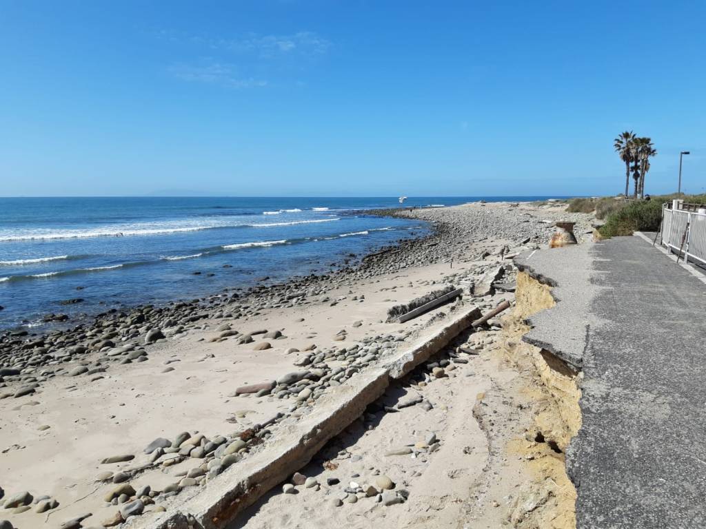 Photograph of crumbling highway behind barricade at a sandy and rocky beach with waves and palm trees in the distance.